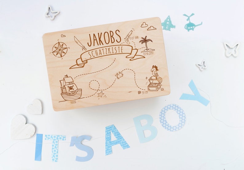 May include: A wooden treasure chest with a pirate theme. The lid is decorated with a map, a pirate ship, a treasure chest, a parrot, and the text "Jakobs Schatzkiste 01.10.2017". The chest is surrounded by blue and white decorations, including the letters "IT'S A BOY".