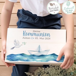 Könnte beinhalten: Eine weiße Holzkiste mit einem blauen Aquarell-Design einer Taube mit einem Olivenzweig und einem Papierboot auf dem Wasser. Die Kiste trägt den Text "Meine Kommunion Anton 05. Mai 2024" auf der Vorderseite.