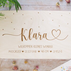 May include: A wooden plaque with the name "Klara" engraved in cursive script, along with a heart symbol and the text "Willkommen kleines Wunder" (Welcome little miracle). The plaque also includes the date "14.01.2022", time "12:22 Uhr", length "46 CM", and weight "3.452 G".