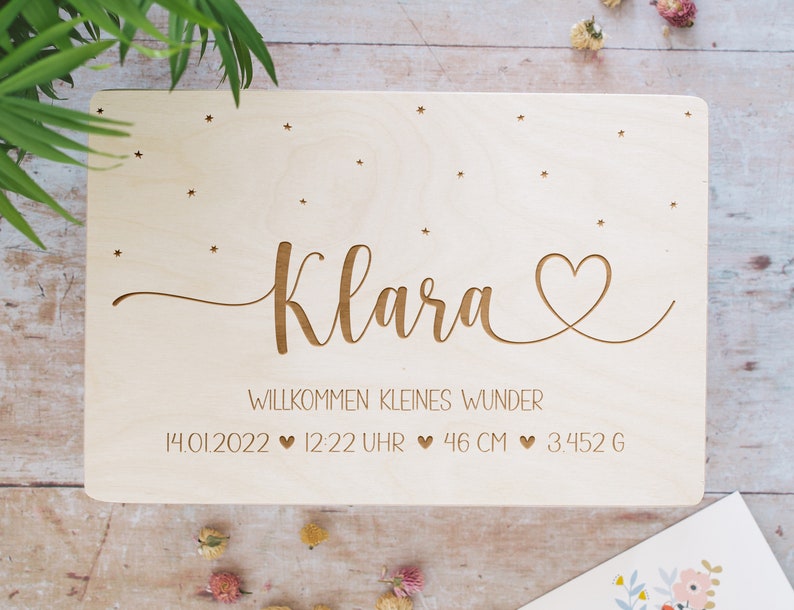 May include: A wooden plaque with the name "Klara" engraved in cursive script, along with a heart symbol and the text "Willkommen kleines Wunder" (Welcome little miracle). The plaque also includes the date "14.01.2022", time "12:22 Uhr", length "46 CM", and weight "3.452 G".