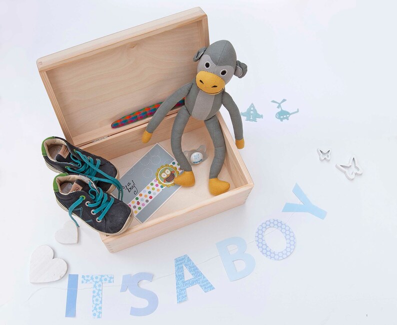 May include: A wooden box with a gray stuffed monkey toy inside. The box is open and there are baby shoes and a card that says "It's a boy!" on the inside. The box is surrounded by blue and white paper cutouts that spell out "It's a boy!"