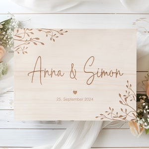 Könnte beinhalten: Eine Holzkiste mit einer hellen Holzoptik und gravierten Blumendesigns. Die Kiste ist personalisiert mit den Namen "Anna & Simon" und dem Datum "25. September 2024".