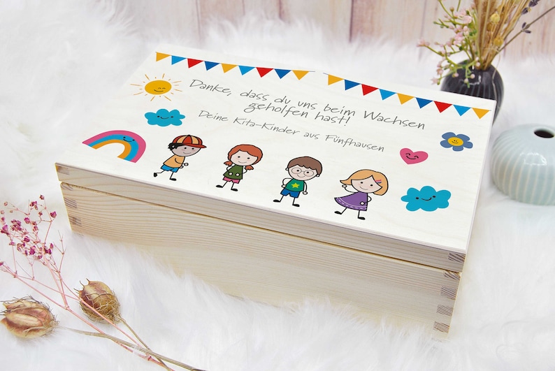 K&ouml;nnte beinhalten: Eine Holzkiste mit einem wei&szlig;en Deckel, der mit bunten Zeichnungen von Kindern, einem Regenbogen, einer Sonne und Wolken verziert ist. Der Deckel tr&auml;gt den Text "Danke, dass du uns beim Wachsen geholfen hast! Deine Kita-Kinder aus F&uuml;nfhausen".