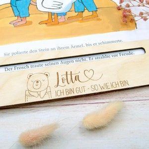 May include: A wooden bookmark with a cutout window, featuring a bear illustration and the text "Lotta ICH BIN GUT - SO WIE ICH BIN". The bookmark is placed on an open book with text visible through the window.