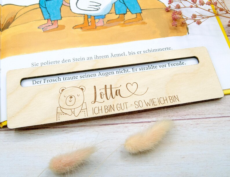 May include: A wooden bookmark with a cutout window, featuring a bear illustration and the text "Lotta ICH BIN GUT - SO WIE ICH BIN". The bookmark is placed on an open book with text visible through the window.
