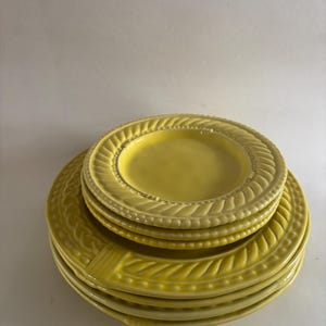 Puede incluir: Una pila de platos de cer&aacute;mica amarillos. Los platos tienen un borde decorativo en forma de cuerda. La pila incluye tanto platos grandes como platos m&aacute;s peque&ntilde;os, todos en un color amarillo soleado a juego. Los platos est&aacute;n apilados sobre una superficie blanca.