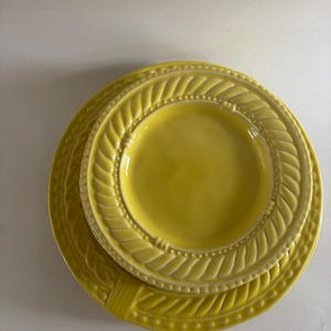 Puede incluir: Una pila de tres platos de cer&aacute;mica amarillos. El plato m&aacute;s grande tiene un borde decorativo en forma de cuerda. El plato del medio es m&aacute;s peque&ntilde;o y tiene un dise&ntilde;o similar. El plato m&aacute;s peque&ntilde;o es liso. Los platos son de un amarillo brillante.