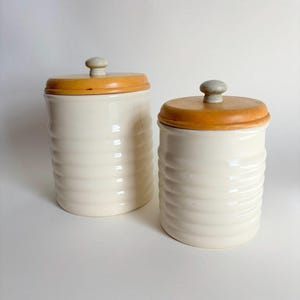 Puede incluir: Dos botes de cerámica blanquecina con laterales acanalados y tapas de madera. Las tapas son de color marrón claro y están rematadas con un pequeño pomo redondo gris. Un artículo de almacenamiento de cocina.