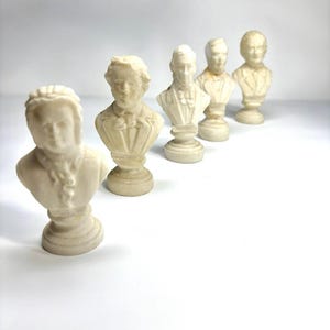 May include: Five off-white, miniature busts of historical figures are arranged in a diagonal line. Each bust features a detailed depiction of a person's head and shoulders, mounted on a pedestal base. The busts are made of a light-colored material, possibly plaster or ceramic.