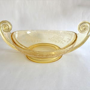 May include: A vintage amber-colored glass bowl with a decorative design. The bowl has a rounded base and two ornate, scrolled handles. The interior features an etched pattern, and the overall design suggests a classic, elegant aesthetic.