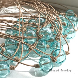May include: A close-up of a pile of turquoise glass bead necklaces with brown leather cord. The necklaces are arranged in a circular pattern, with the beads overlapping each other. The text "Sand Kissed Jewelry" is visible at the bottom of the image.