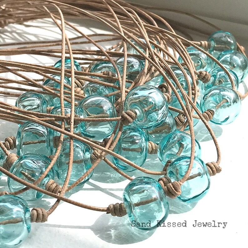 May include: A close-up of a pile of turquoise glass bead necklaces with brown leather cord. The necklaces are arranged in a circular pattern, with the beads overlapping each other. The text "Sand Kissed Jewelry" is visible at the bottom of the image.