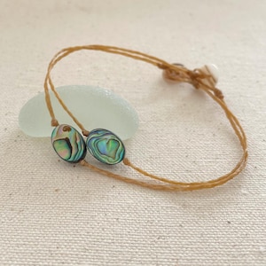 May include: A brown cord bracelet with two oval-shaped abalone shell beads. The beads have iridescent green, blue, and purple colors.