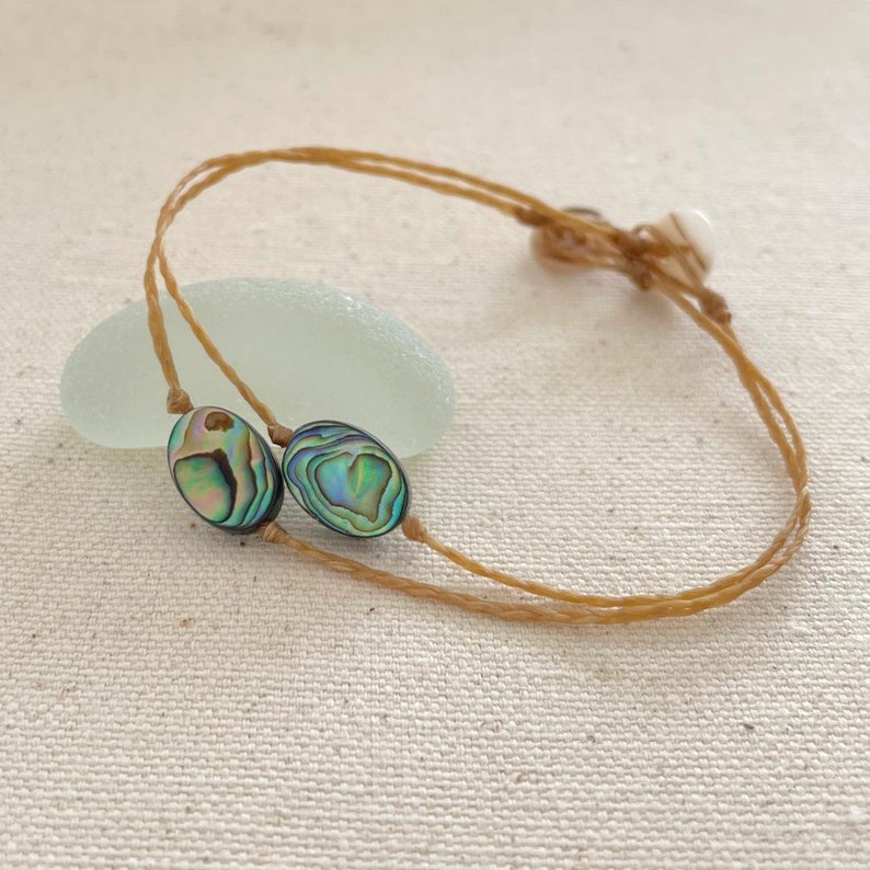 May include: A brown cord bracelet with two oval-shaped abalone shell beads. The beads have iridescent green, blue, and purple colors.