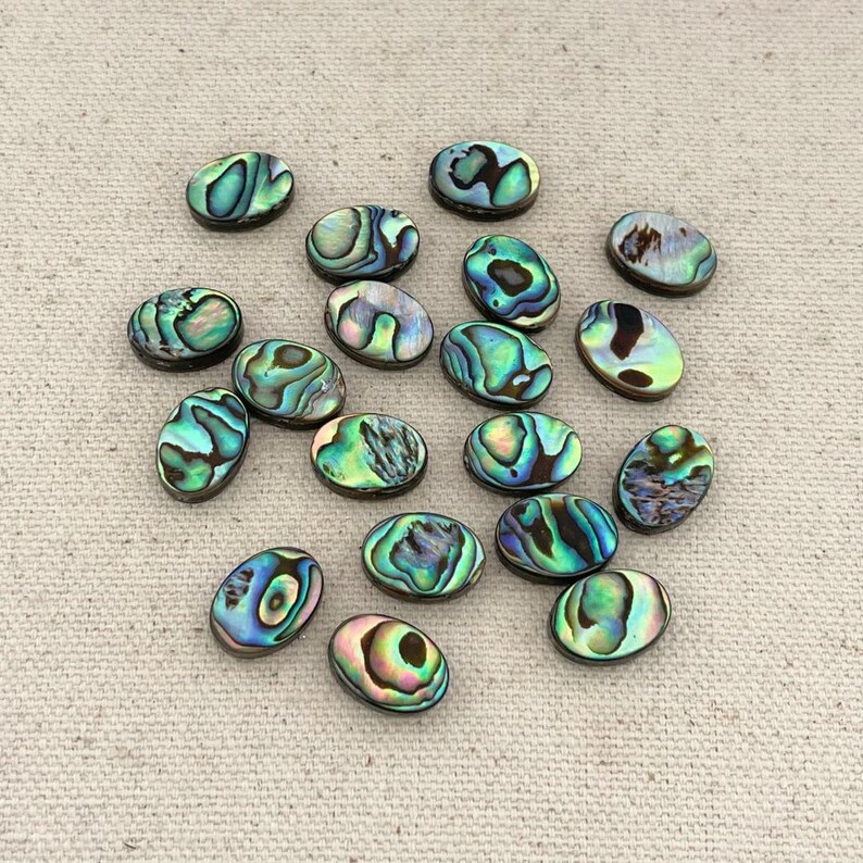 May include: A collection of 17 oval-shaped abalone shell cabochons in various shades of green, blue, and brown. The cabochons are iridescent and have a natural, organic look.