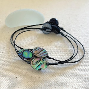 May include: A bracelet with three iridescent abalone shell beads and black cord. The bracelet is tied with a knot and has a simple design.