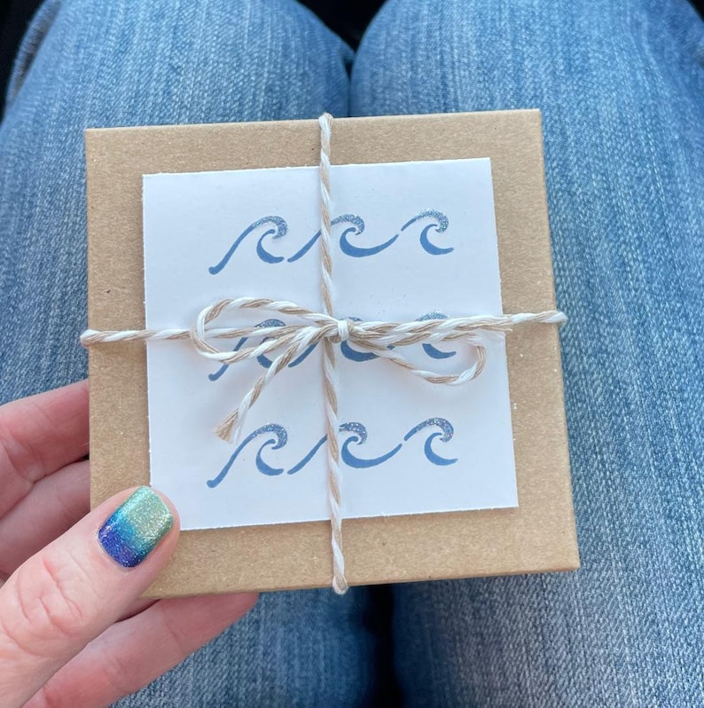 May include: A small, brown gift box tied with twine and a white bow. The box has a white card attached to the front with a blue wave design.
