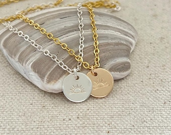 May include: Two delicate necklaces with a sunrise design pendant. One necklace has a silver chain and pendant, the other has a gold chain and pendant. Both pendants are round and feature a simple sunrise design.