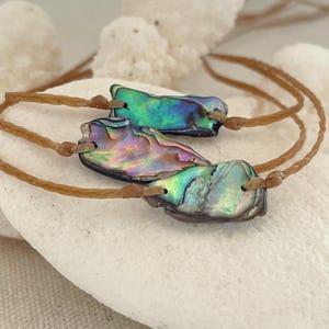 May include: Three bracelets featuring irregular, iridescent abalone shell pieces with vibrant green, blue, and purple hues. Each shell segment is drilled and secured onto a thin, braided tan cord, creating a set of adjustable jewelry pieces.