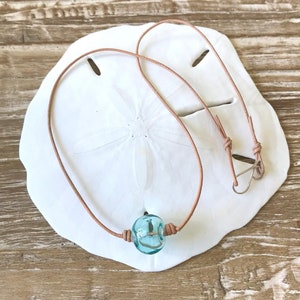 May include: A light blue glass bead necklace with a brown leather cord. The necklace is displayed on a white sand dollar.