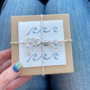 Op de afbeelding: Een klein, bruin cadeaudoosje, vastgebonden met touw en een witte strik. Het doosje heeft een witte kaart die aan de voorkant is bevestigd met een blauw golvend ontwerp.