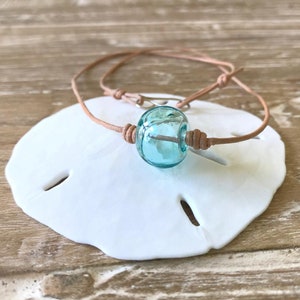 May include: A turquoise glass bead necklace with a brown leather cord. The necklace is displayed on a white seashell.