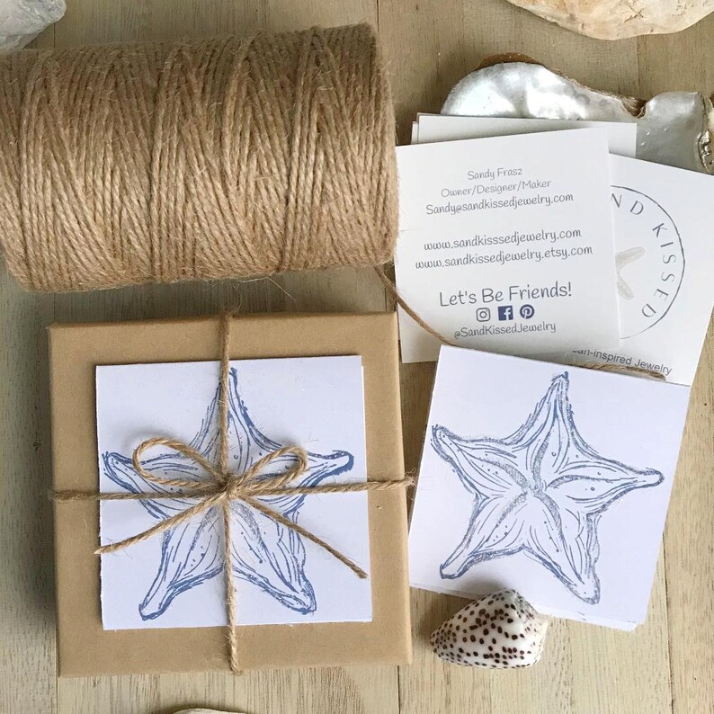 May include: A brown gift box tied with twine and a blue and white starfish illustration on the top. The box is sitting on a wooden surface with a business card that says "Sandy Kissed Jewelry" and "Let's Be Friends!" with social media icons.