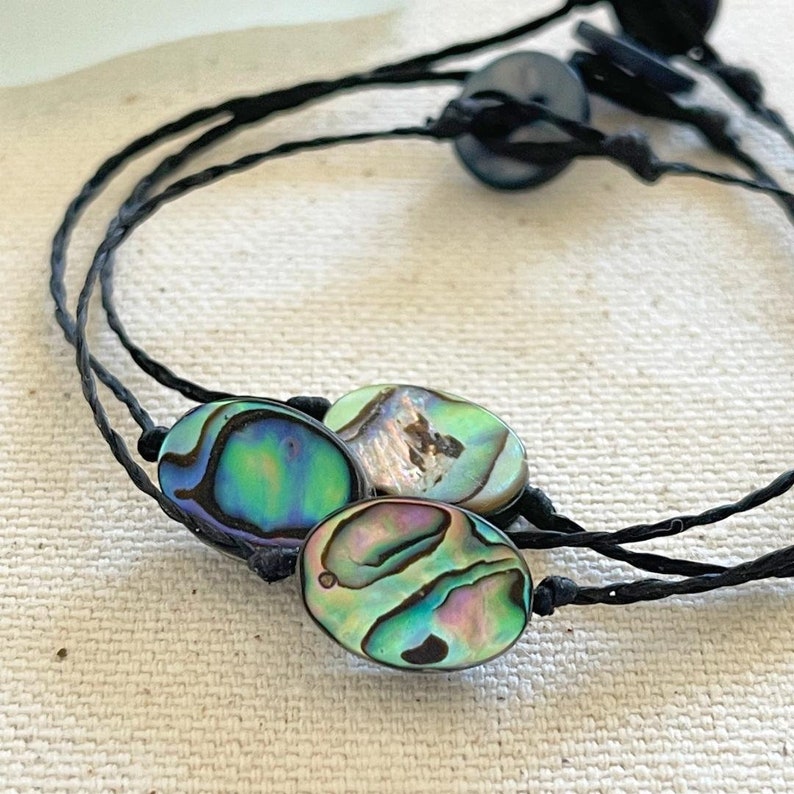 May include: Three oval-shaped abalone shell charms on a black cord bracelet. The shells have iridescent green, blue, and purple colors.