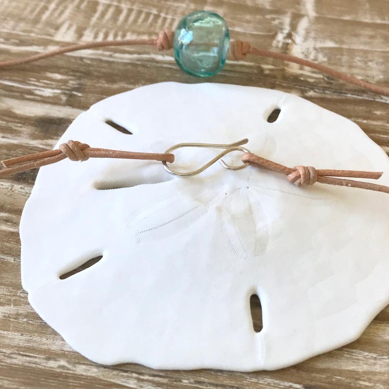 May include: A white sand dollar with a light brown leather cord and a turquoise glass bead. The cord has a silver clasp.