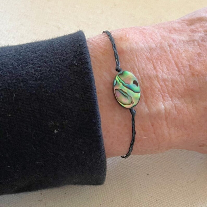 Op de afbeelding: Een armband van zwart koord met een ronde hanger van abalone-schelp. De schelp heeft iriserende groene, blauwe en paarse kleuren.