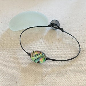 Op de afbeelding: Een armband van zwart koord met een groene, blauwe en iriserende abalone-schelpbedel en een zwarte knoopsluiting.