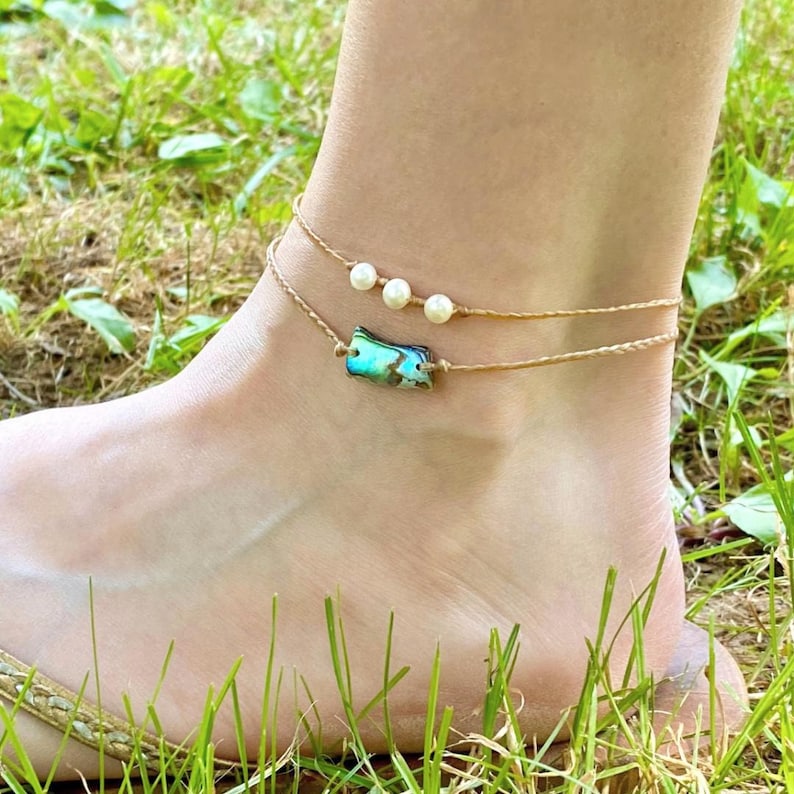 May include: A tan ankle bracelet with a small abalone shell charm and three white pearl beads.
