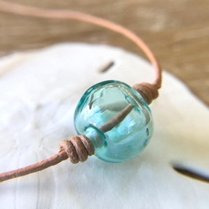 May include: A light blue glass bead on a brown leather cord. The bead is round and has a smooth, polished surface.