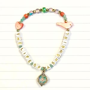 Op de afbeelding: Een kralenarmband met de tekst "THE LIFE OF A SHOWGIRL". De armband heeft witte letterkralen, roze hart- en vogelbedels en een goudkleurige hanger met een kleurrijke edelsteen. De kralen zijn in tinten roze, groen en goud.