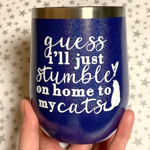 Puede incluir: Un vaso de acero inoxidable azul oscuro con un acabado de brillo blanco. El vaso tiene un diseño de vinilo blanco que dice "guess I'll just stumble on home to my cats" con una silueta blanca de un gato.