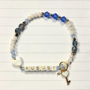 May include: A white and blue beaded bracelet with a gold charm and a white heart charm. The bracelet has a white tile charm with the word "AUGUST" spelled out in gold letters.