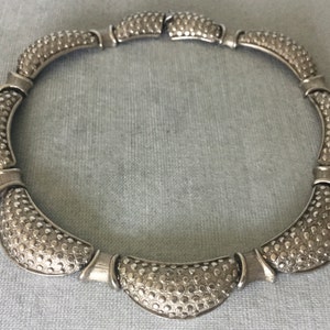 May include: A silver-toned necklace with a textured, dotted pattern. The necklace is composed of curved, segmented pieces connected by smaller, smooth sections. The design is symmetrical and appears to be made of metal.
