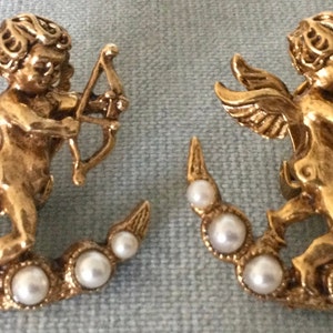Puede incluir: Un par de pendientes de tono dorado con un querubín sosteniendo un arco y una flecha, de pie sobre una luna creciente con detalles de perlas falsas.