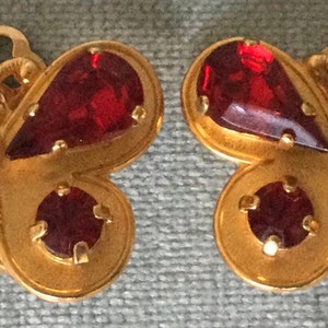 May include: A pair of gold-toned butterfly-shaped earrings. Each earring features a large, teardrop-shaped, red gemstone on the wings, a smaller red gemstone on the body, and a clear, round gemstone in the center. The earrings have a detailed, textured design.
