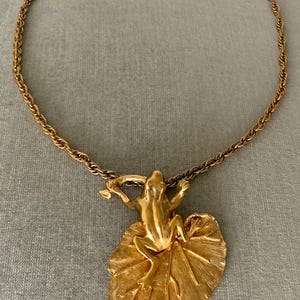 May include: Gold-toned necklace featuring a frog pendant resting on a leaf. The frog is detailed with outstretched arms, and the leaf has textured veins. The chain is a twisted rope design, creating a vintage aesthetic.