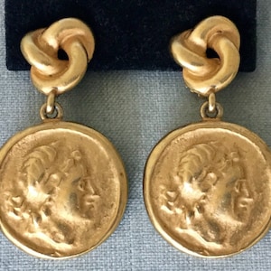 Pode incluir: Um par de brincos dourados. Cada brinco apresenta uma parte superior em forma de nó e um pendente circular com um relevo de perfil. Os pendentes retratam uma figura clássica. Os brincos são exibidos sobre um fundo escuro.
