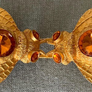 Op de afbeelding: Een goudkleurige metalen broche in de vorm van twee gestileerde bijen die tegenover elkaar staan. Elke bij heeft grote, facetgeslepen amberkleurige kristallen ogen en kleinere kristallen. De vleugels en lichamen hebben gedetailleerde texturen.
