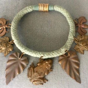May include: A light green rope necklace with bronze-colored charms. The charms include a frog, leaves, grapes, and bees. The necklace has gold-colored accents and a circular design.