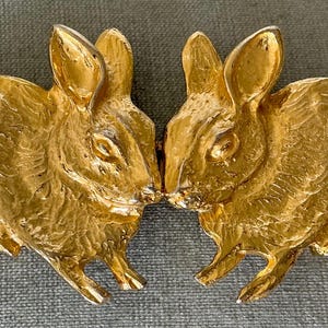 Puede incluir: Una hebilla de cinturón de metal dorado con dos conejos frente a frente. Los conejos están representados de perfil con detalles de pelaje texturizado, orejas puntiagudas y cuerpos redondeados. Las dos mitades se unen en el centro.