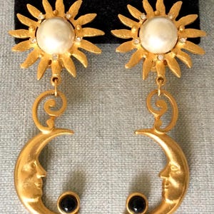 May include: Gold tone earrings with a sun and moon design. The sun features a large pearl and smaller rhinestones. The moon has a black stone at the bottom.