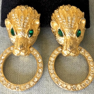 Op de afbeelding: Gouden oorknoppen met een leeuwenkop, groene edelsteenogen en een ring van helder kristal.