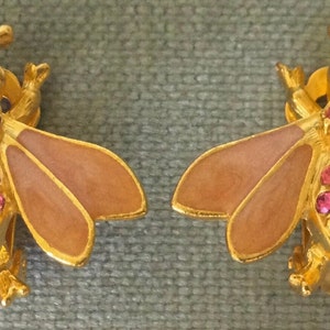 May include: A pair of gold-toned bee-shaped earrings. The earrings feature pink crystal accents on the body and light brown enamel wings. The bee's body is textured, and the wings are outlined in gold.