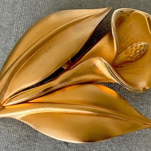 May include: A gold-coloured metal brooch shaped like a calla lily with two leaves. The brooch has a smooth, polished finish. The flower's centre detail is visible. It is set against a grey background.