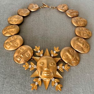 May include: A gold-toned statement necklace featuring a sunburst pendant with a face and clear crystal accents. The necklace includes oval-shaped links with embossed faces. The necklace is a bold, artistic piece of jewelry.