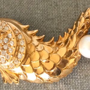 Puede incluir: Broche de pez dorado con una perla en la boca y otra en la cola. El pez está incrustado con cristales transparentes y tiene un ojo de gema roja. La cola tiene forma de corazón y también está incrustada con cristales.